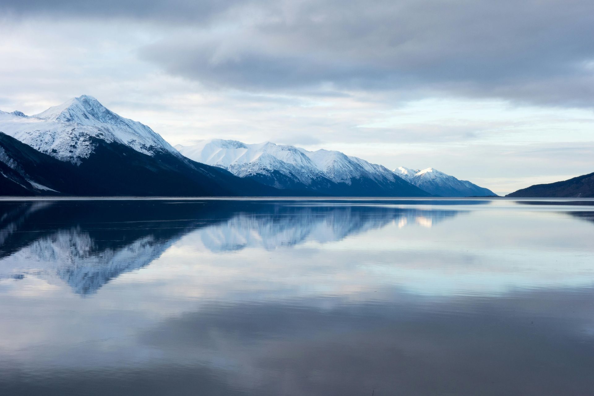Snow-capped mountains reflect in calm, blue water under a cloudy sky.