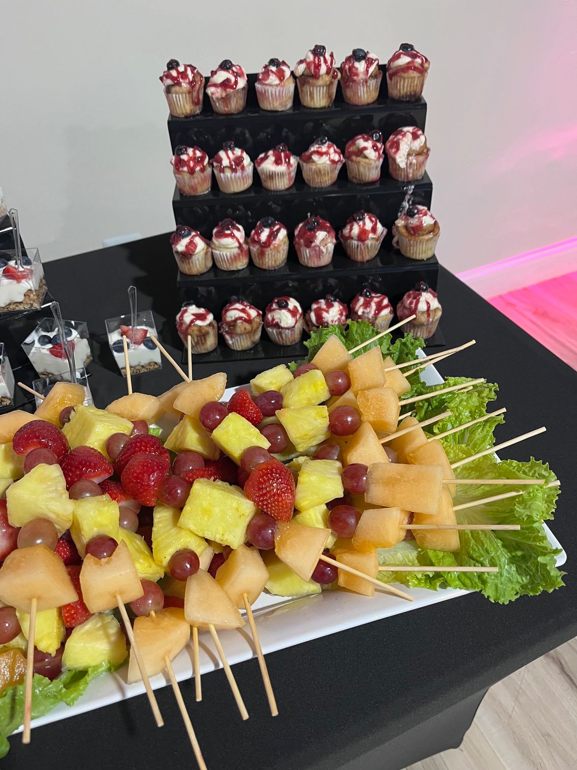 Dessert display: Fruit skewers, cupcakes with berry topping, black table, white platter.
