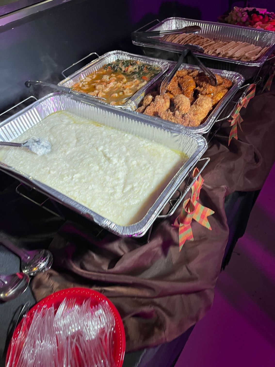 Buffet with grits, collard greens, fried chicken, and meat. Red plate with plastic cutlery.