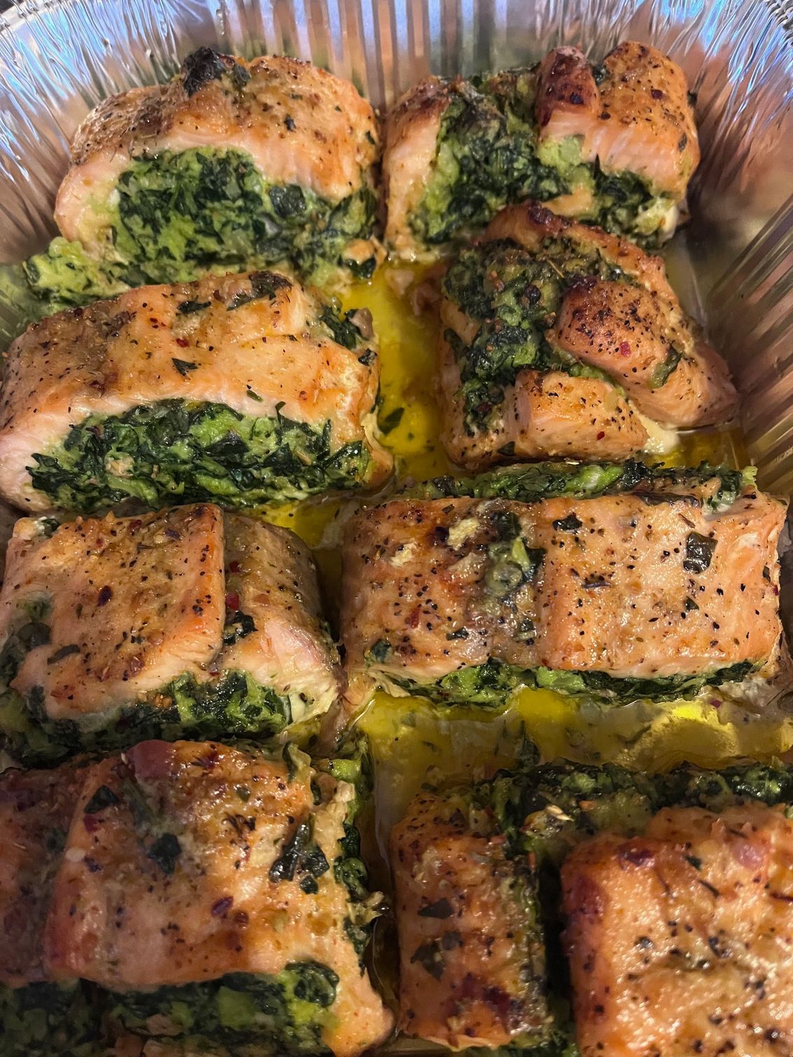 Stuffed salmon fillets with green filling in a foil pan, drizzled with sauce, and seasoning.