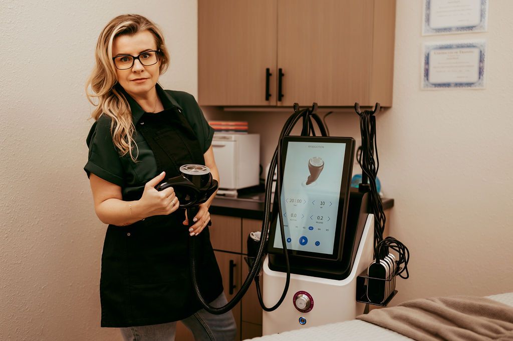 Woman with glasses and a dark apron holding a device, standing near medical equipment in a clinic.