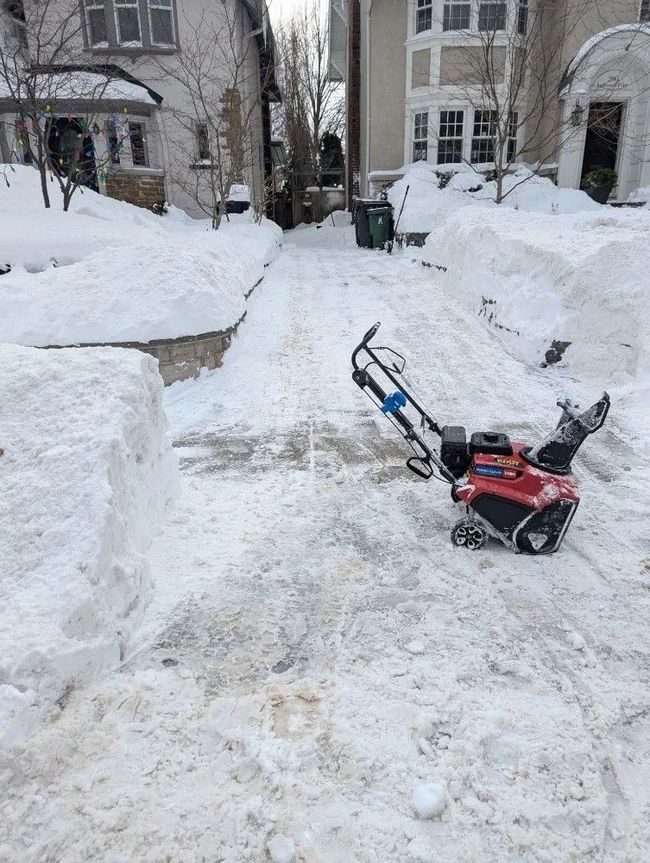 Snow-cleared walkway between tall snowbanks in front of apartment buildings, with a red snow blower in the foreground