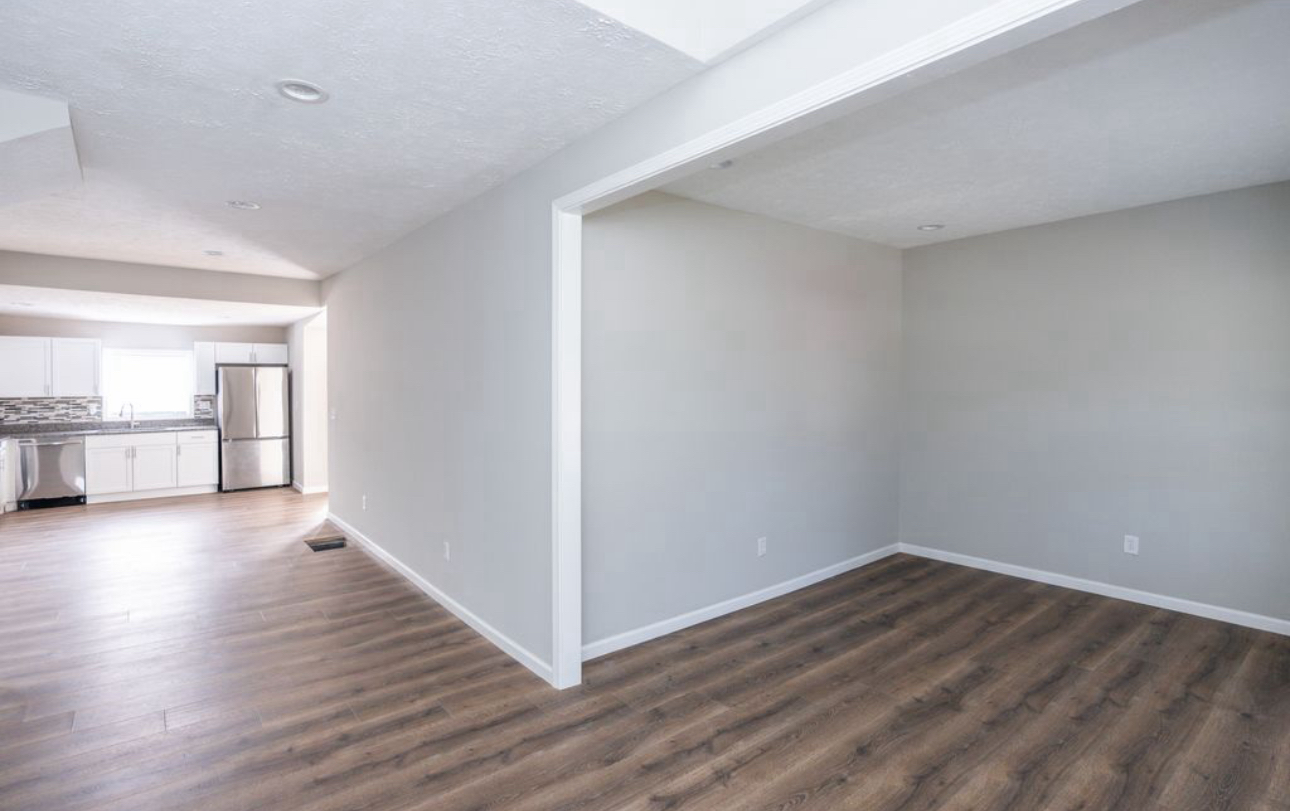 An empty room with hardwood floors and a kitchen in the background.