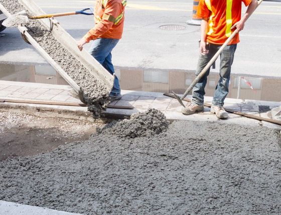 Two construction workers in orange vests pour wet concrete from a chute onto a sidewalk.