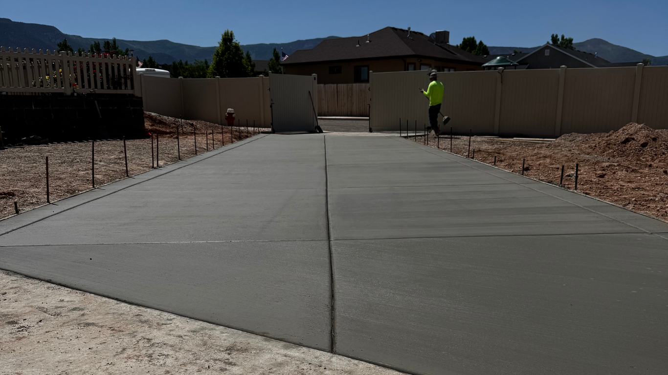 Freshly poured concrete driveway with smooth surface, bordered by wooden stakes, leading to a fenced yard.