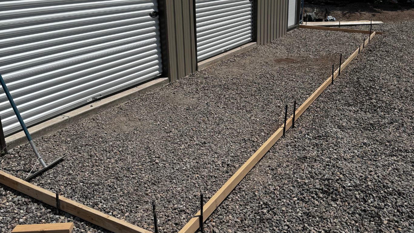 Construction site with gravel ground and wooden frames outlining a foundation area next to a building.