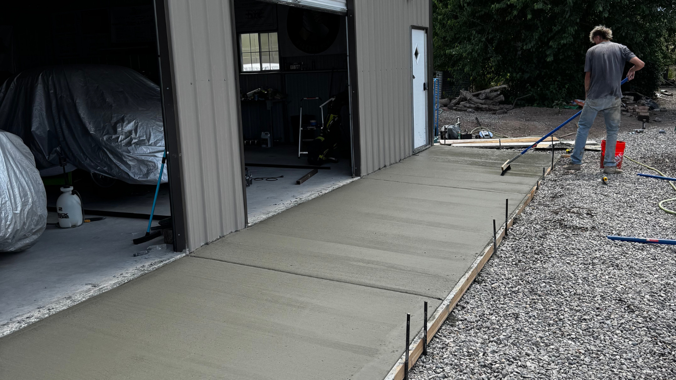 A person smooths freshly poured concrete beside a metal garage, which holds covered vehicles. 