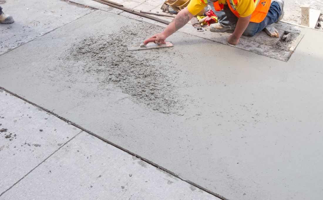 A worker in a yellow vest kneels and smooths wet concrete with a trowel on a city sidewalk, creating an even surface. 