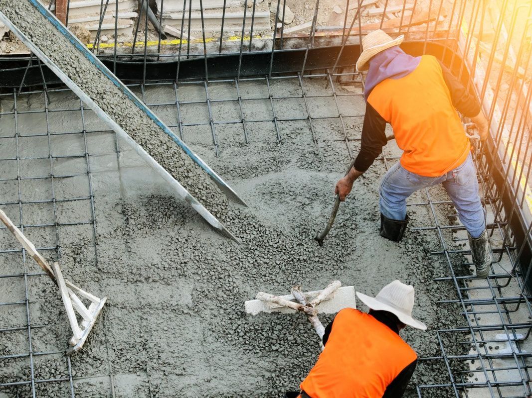 Two construction workers in orange vests and hats spread wet concrete on a steel-reinforced floor using tools. 