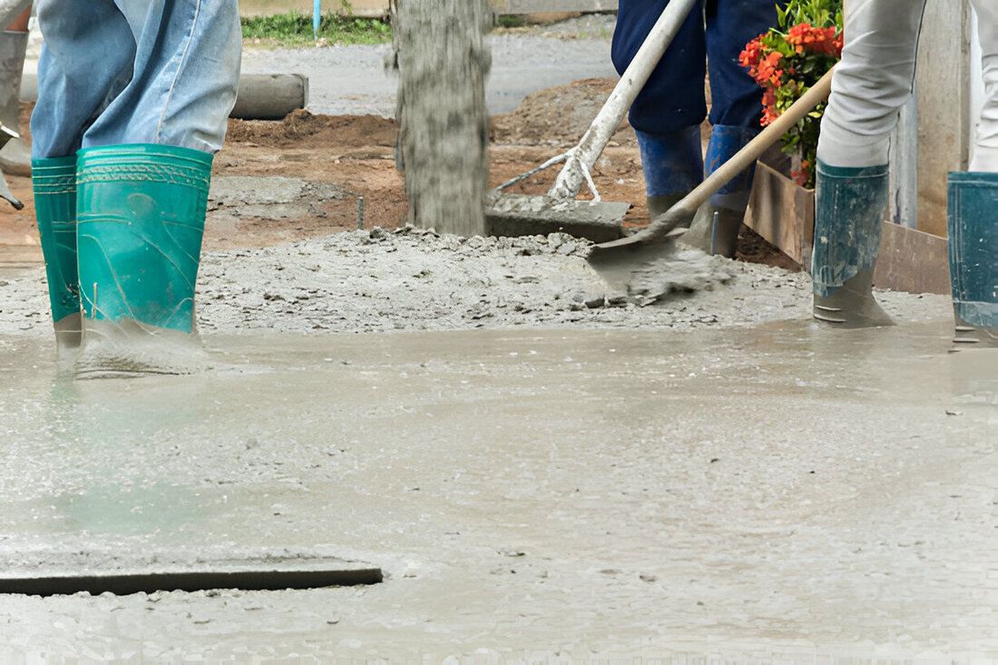 Workers in blue jeans and green boots stand on wet concrete, using tools to smooth it. The scene conveys a sense of teamwork.