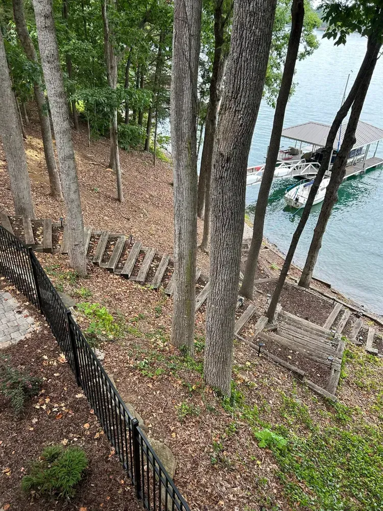 Wooded lakeside slope with stone steps, a black fence, and a dock by calm water