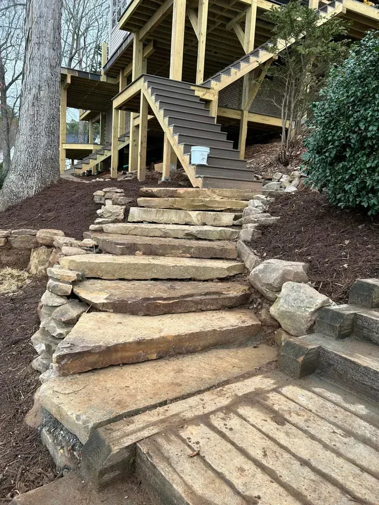 Rustic stone steps leading up to a wooden deck and railing on a hillside amid trees and mulch