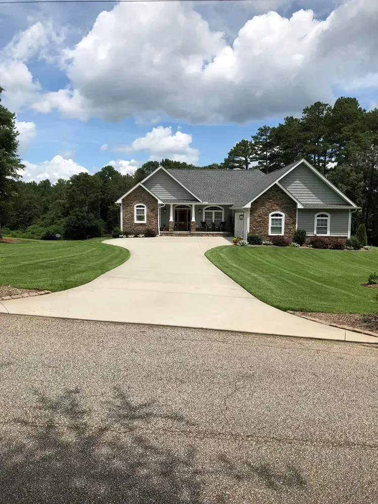 Single-story brick house with a wide driveway, green lawn, and cloudy sky.
