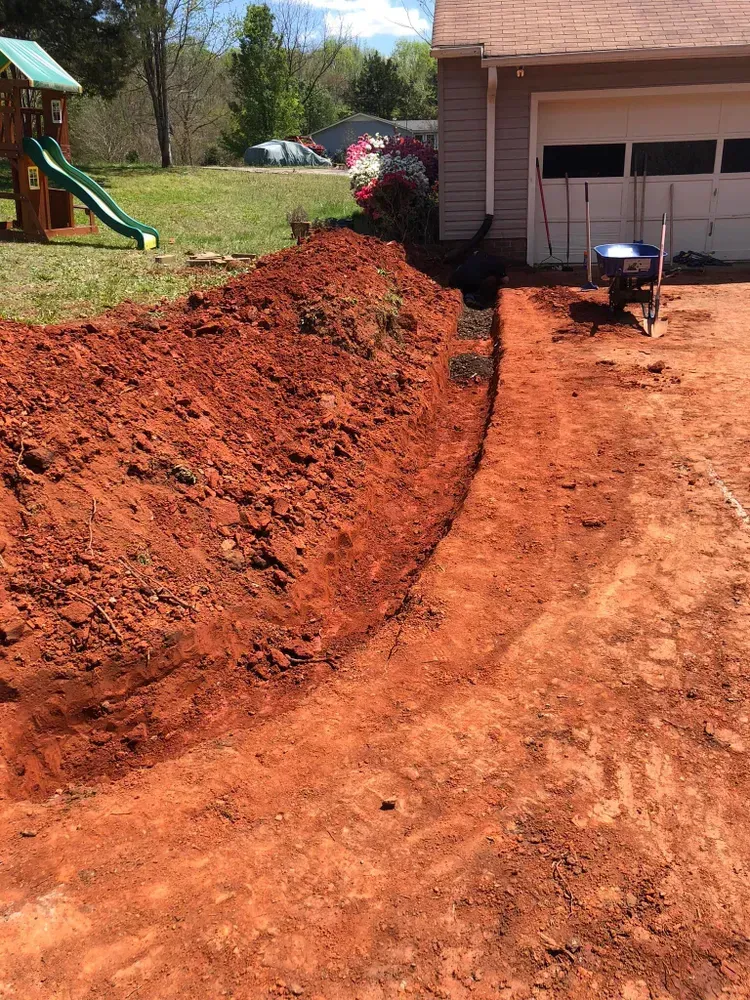 Freshly dug red dirt trench beside a garage, with a wheelbarrow and a slide in the background.