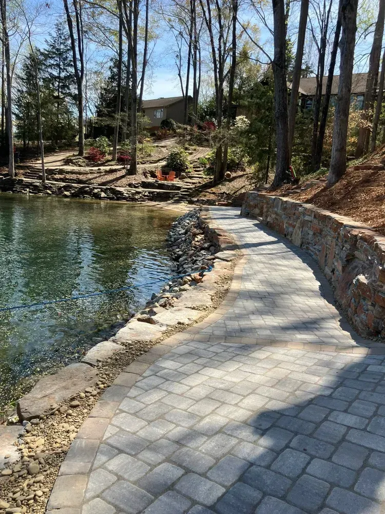 Curved stone walkway along a lakeside, with trees, rocks, and houses in the background.