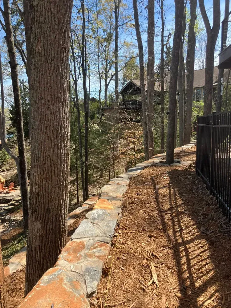 Stone-edged wooded path beside a black fence, with tall trees and a house in the background