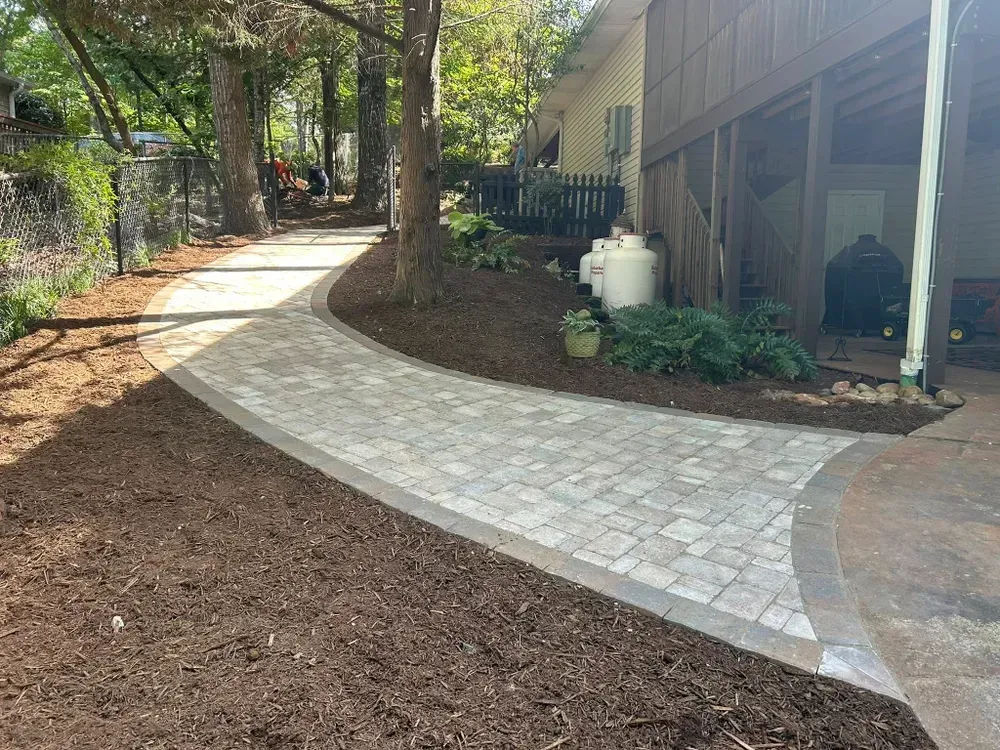 Curved stone walkway through a wooded yard beside a house and patio columns