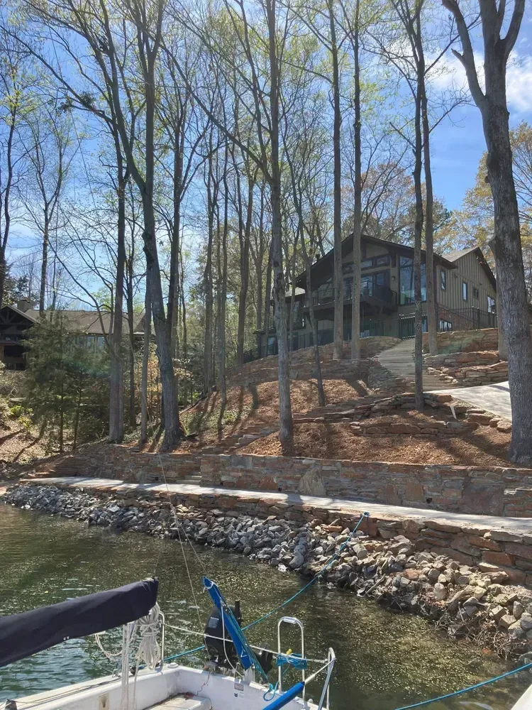Docked boats on a lake beside a stone wall, with a wooded hillside and house in the background.