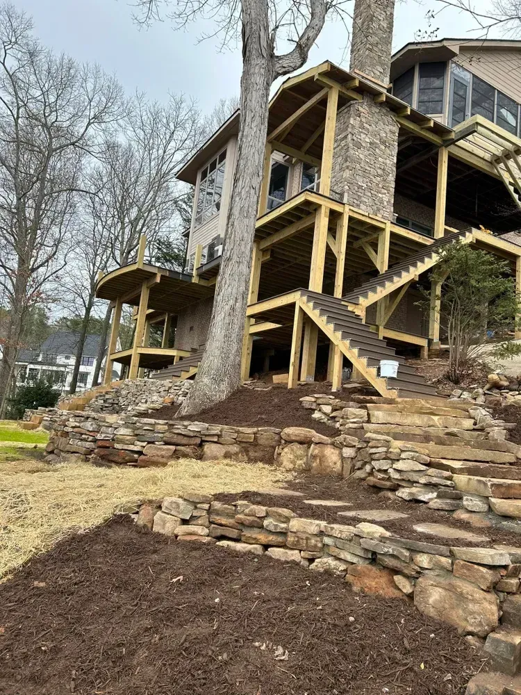 Stone retaining wall and wooden stairs beside a house on a wooded hillside, with bare trees and muddy ground.