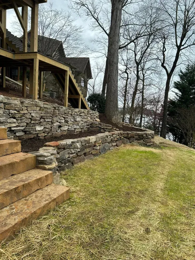 Stone steps and retaining wall beside a grassy slope, with leafless trees and a wooden deck on the left.