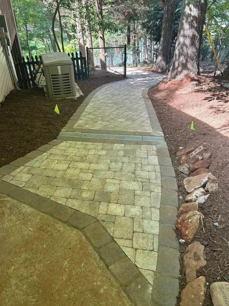 Curved brick walkway through a wooded yard, bordered by dirt and trees, leading to a gate.