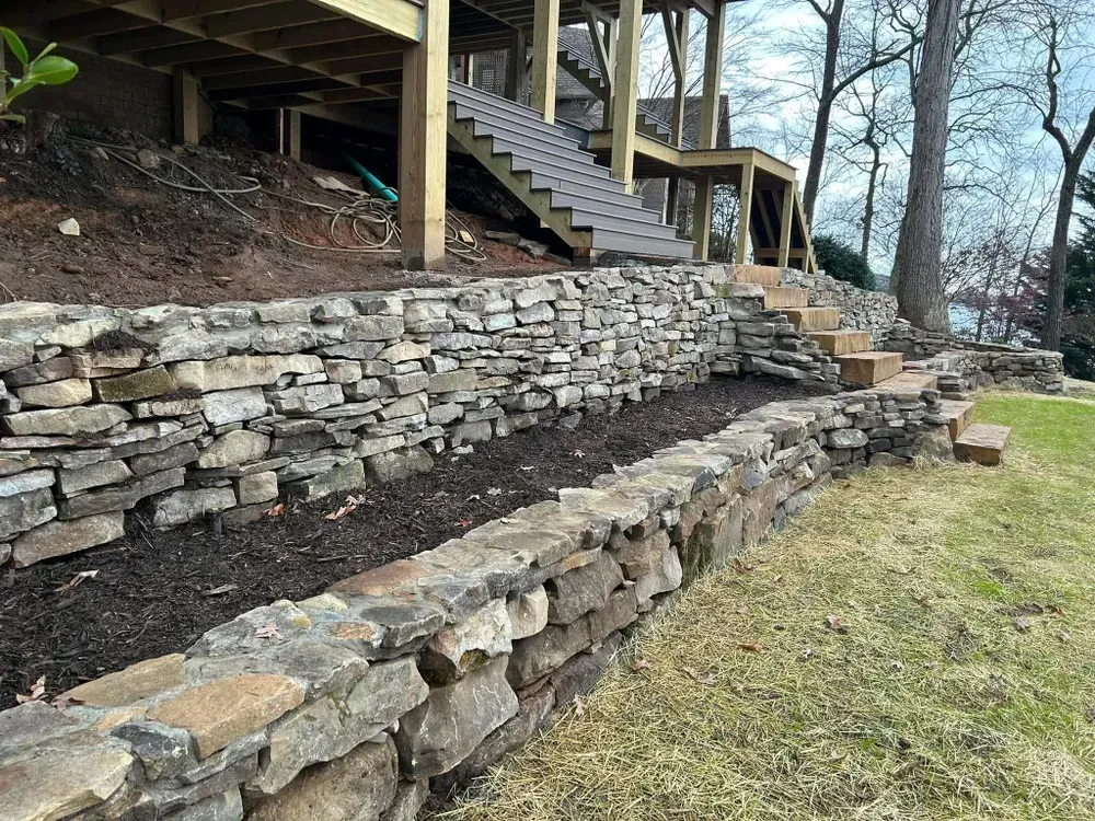 Terraced stone retaining walls under a deck beside outdoor stairs and a grassy yard