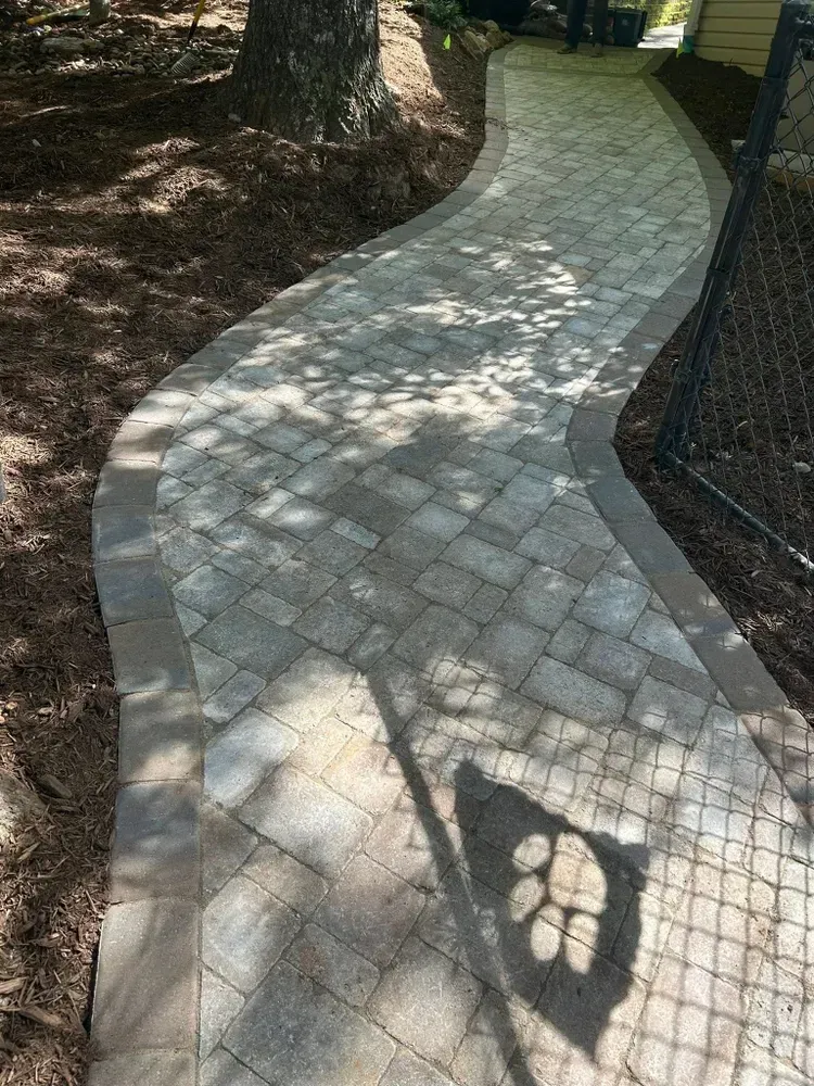 Curved stone walkway through a wooded area, with dappled sunlight and chain-link fence on the right.