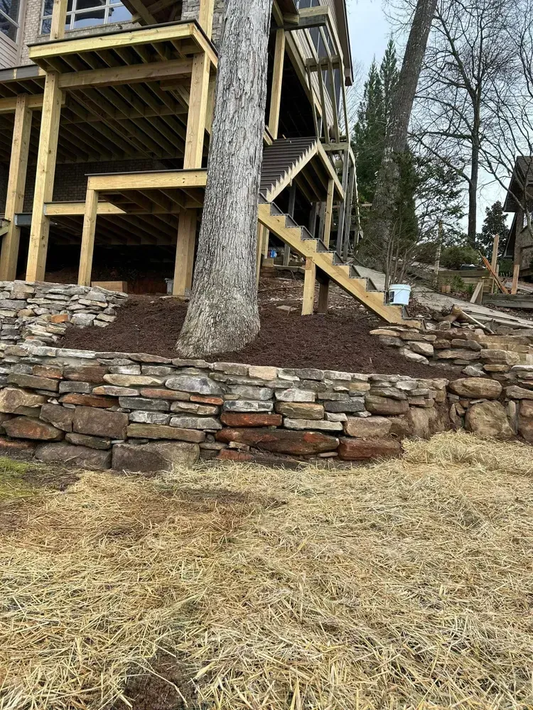 Stone retaining wall and large tree in front of a raised wooden deck with stairs on a wooded slope.
