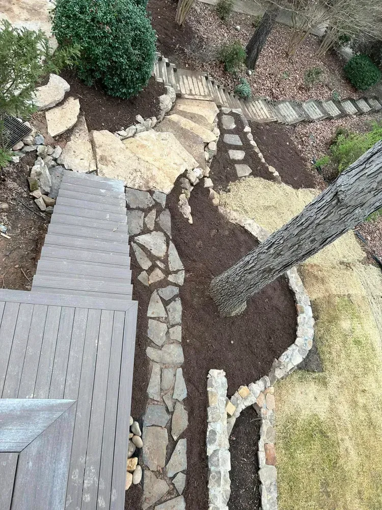 Backyard landscape with stone path, mulch beds, deck edge, and tree trunk