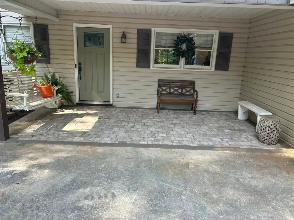 Covered porch with beige siding, green door, bench under a window, and potted plants on a stone patio.