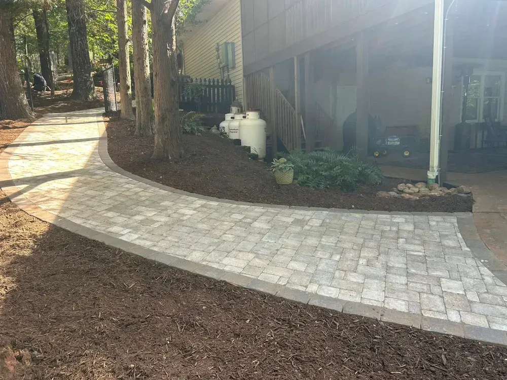 Curved stone walkway beside a house, with plants, mulch, and propane tanks in the yard