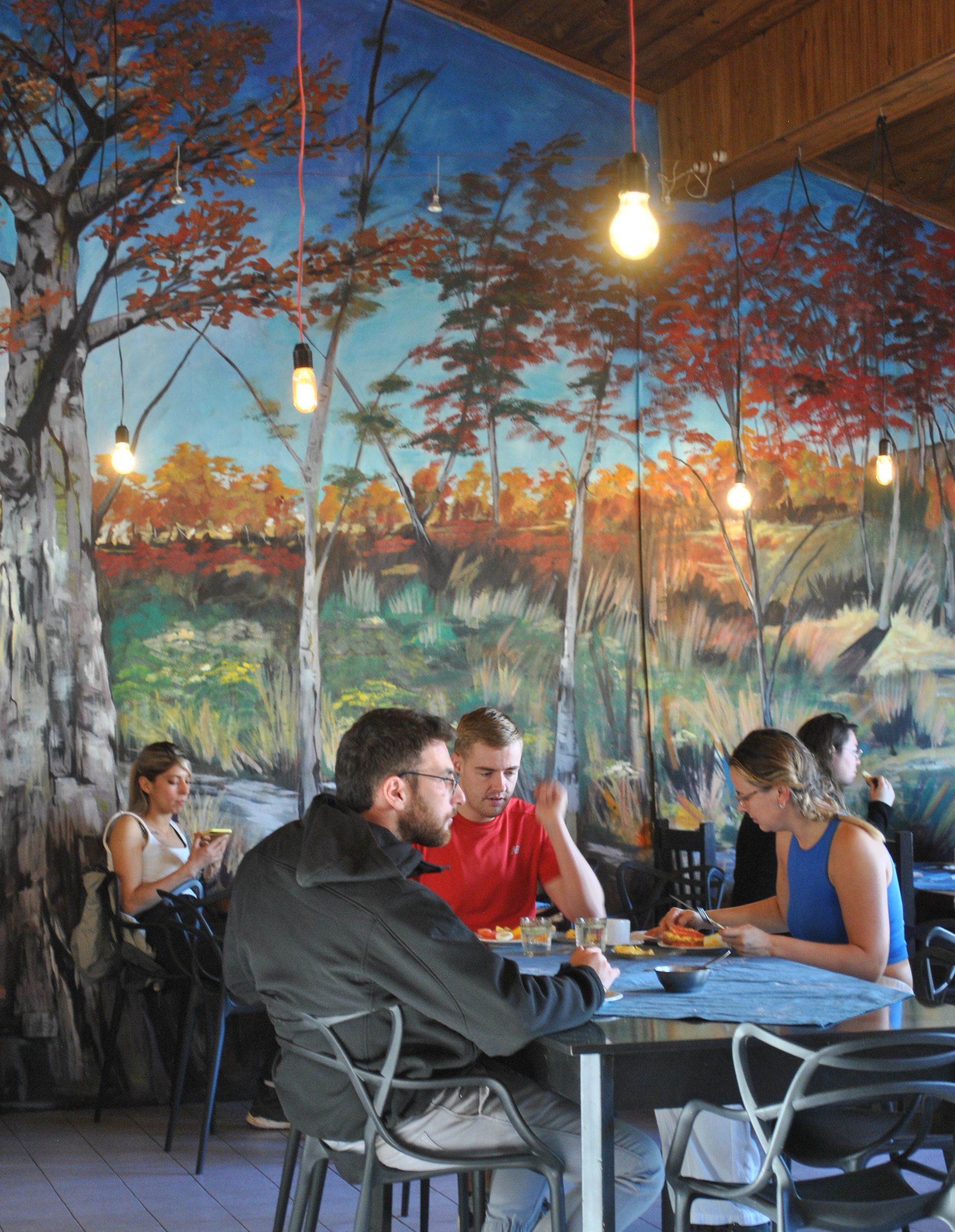 Personas sentadas en mesas dentro de un restaurante con un mural de un bosque en la pared. Lámparas colgantes.
