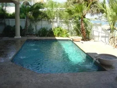 There is a large swimming pool in the backyard of a house surrounded by palm trees.