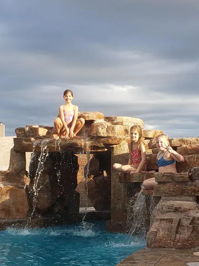 Three young girls are sitting on top of a waterfall next to a pool.