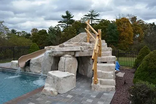 A water slide is built into the rocks next to a swimming pool.