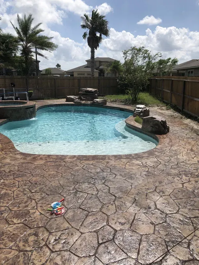 A large swimming pool is surrounded by a stone patio and palm trees.