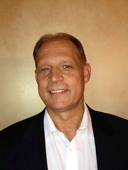 Man in a black blazer and white shirt, smiling. Beige background.