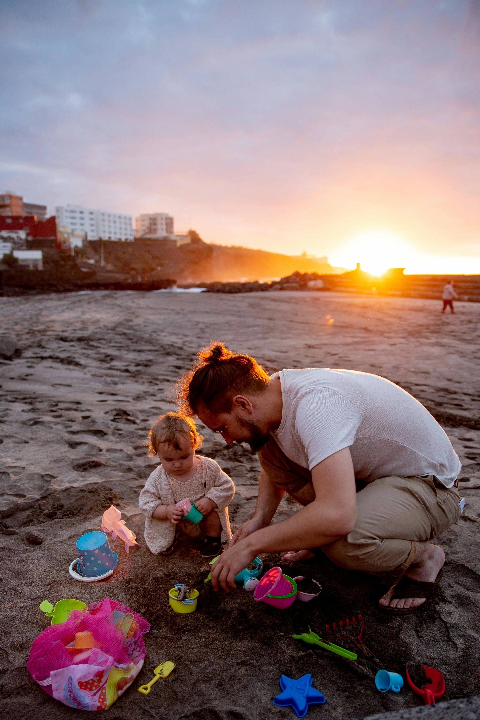 A man and a little girl are playing in the sand on the beach at sunset.