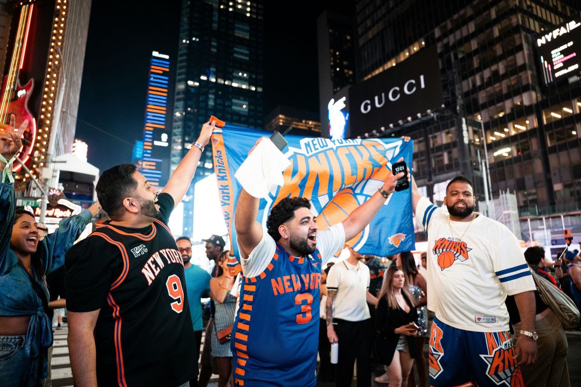 Fans cheering with New York Knicks gear and a blue-and-orange flag in a city square at night