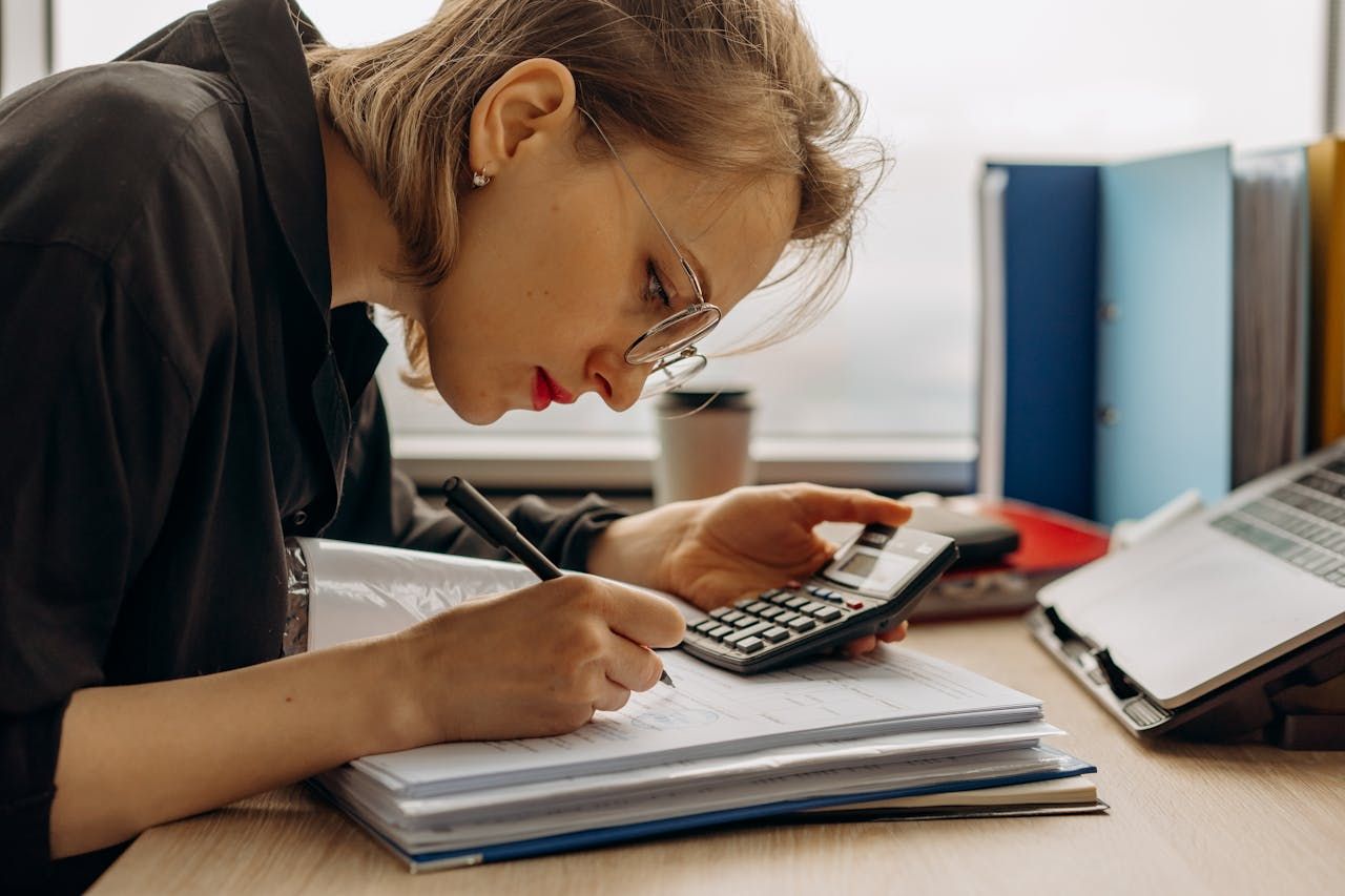 Woman with glasses using a calculator and writing in a notebook at a desk.
