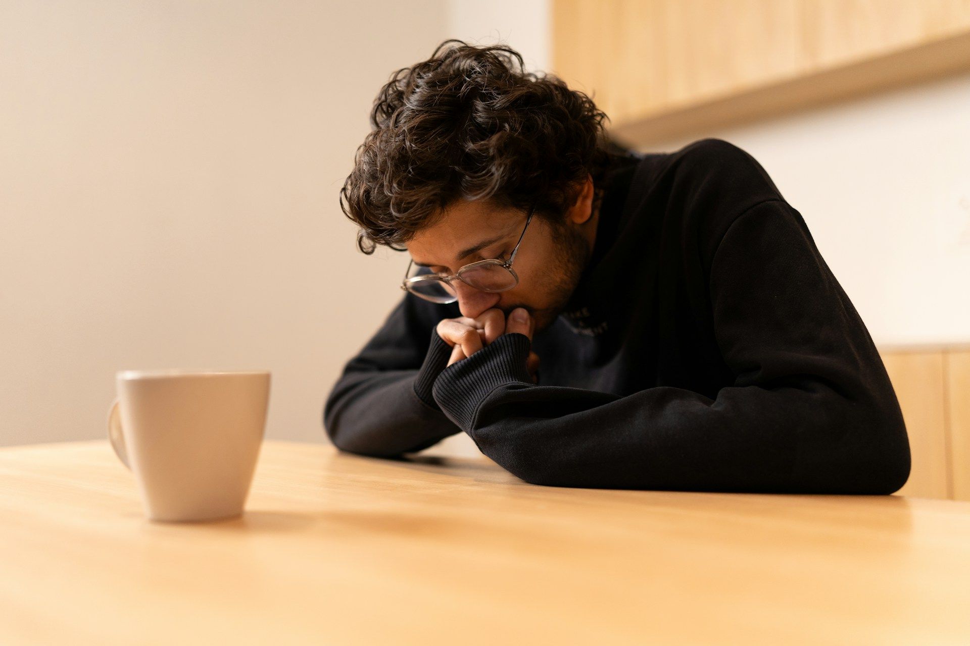 A man, ashamed of a financial mistake, sits at a table, chin resting on folded hands beside a mug.