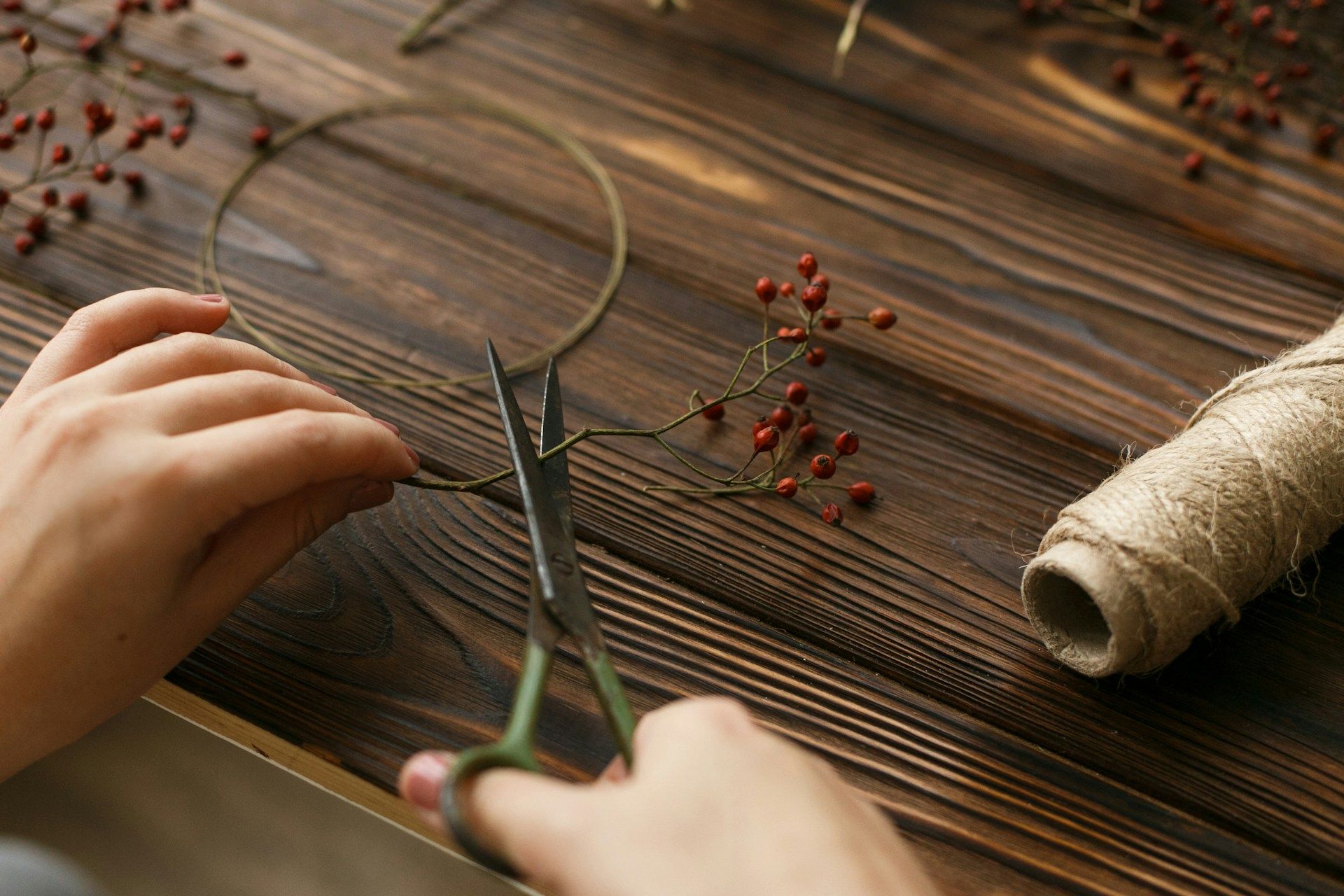 Hands cutting a small branch with berries, making a wreath with scissors on a wood surface.