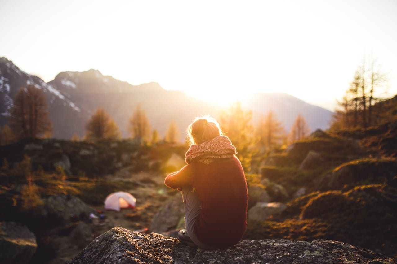 A person is sitting on a rock in the mountains looking at the sun and sky