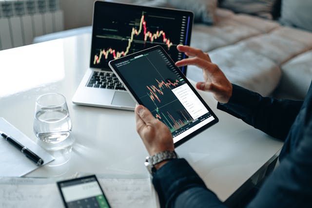 Person analyzing stock charts on a laptop and tablet, in a room.