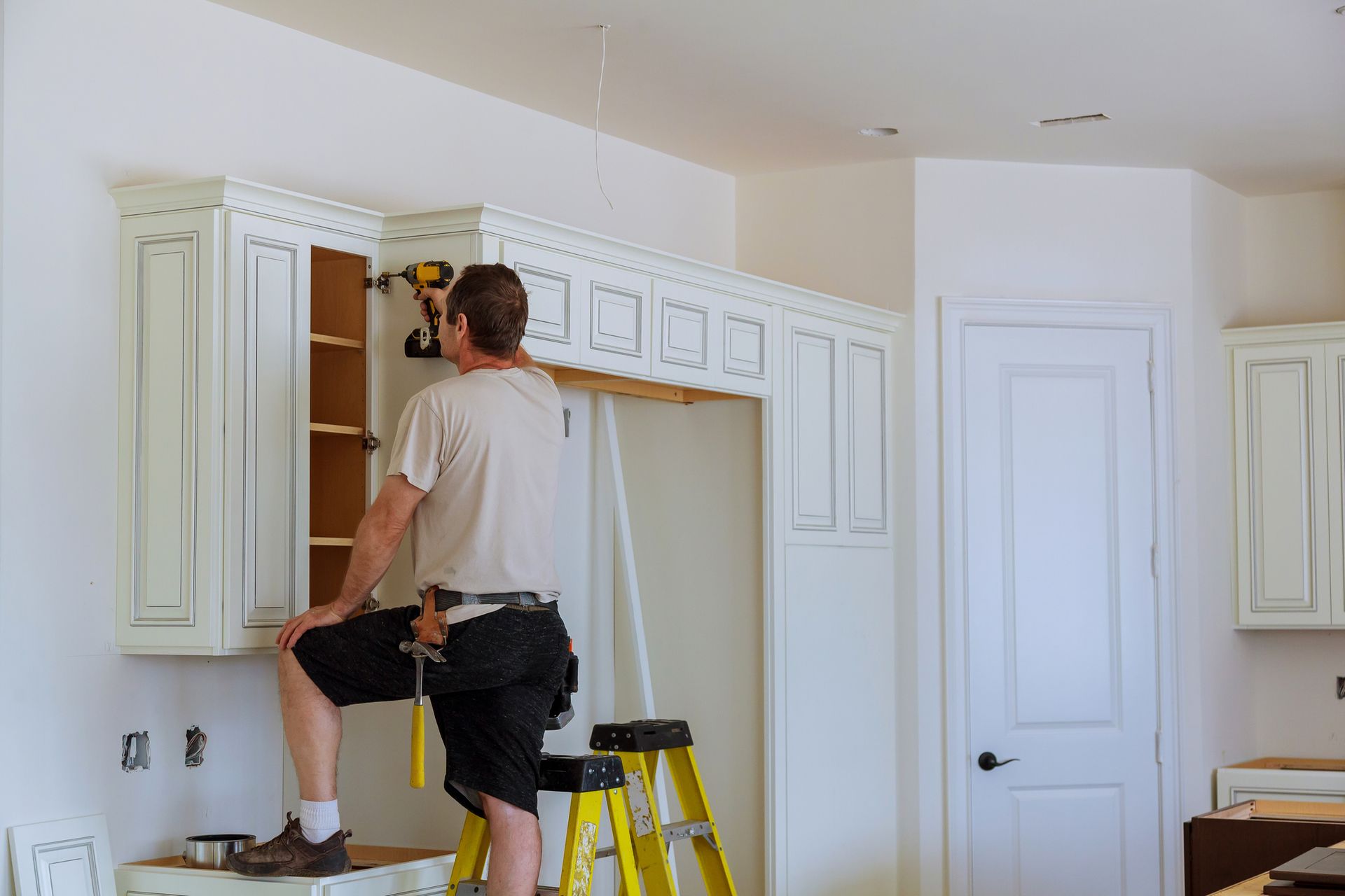 A man is standing on a ladder in a kitchen installing cabinets — Clifton, NJ — New Alp Construction