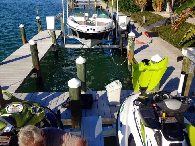 A boat on a lift, with two jet skis docked, on a wooden pier. Bright sunny day.