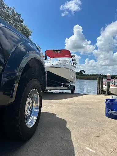 Black truck towing a white boat on a trailer at a boat ramp on a sunny day.