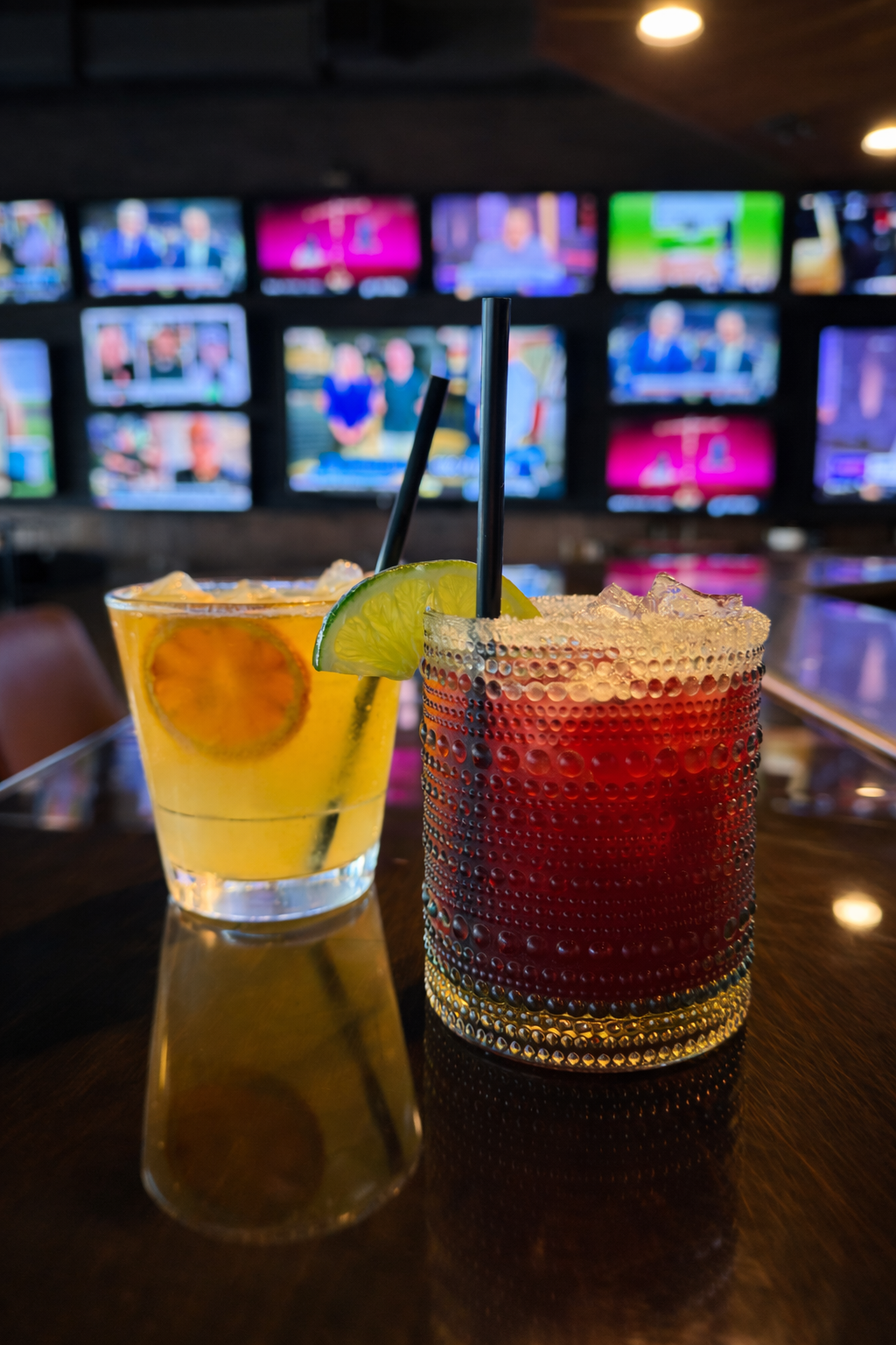 Two cocktails on a dark bar counter with a wall of television screens in the background.