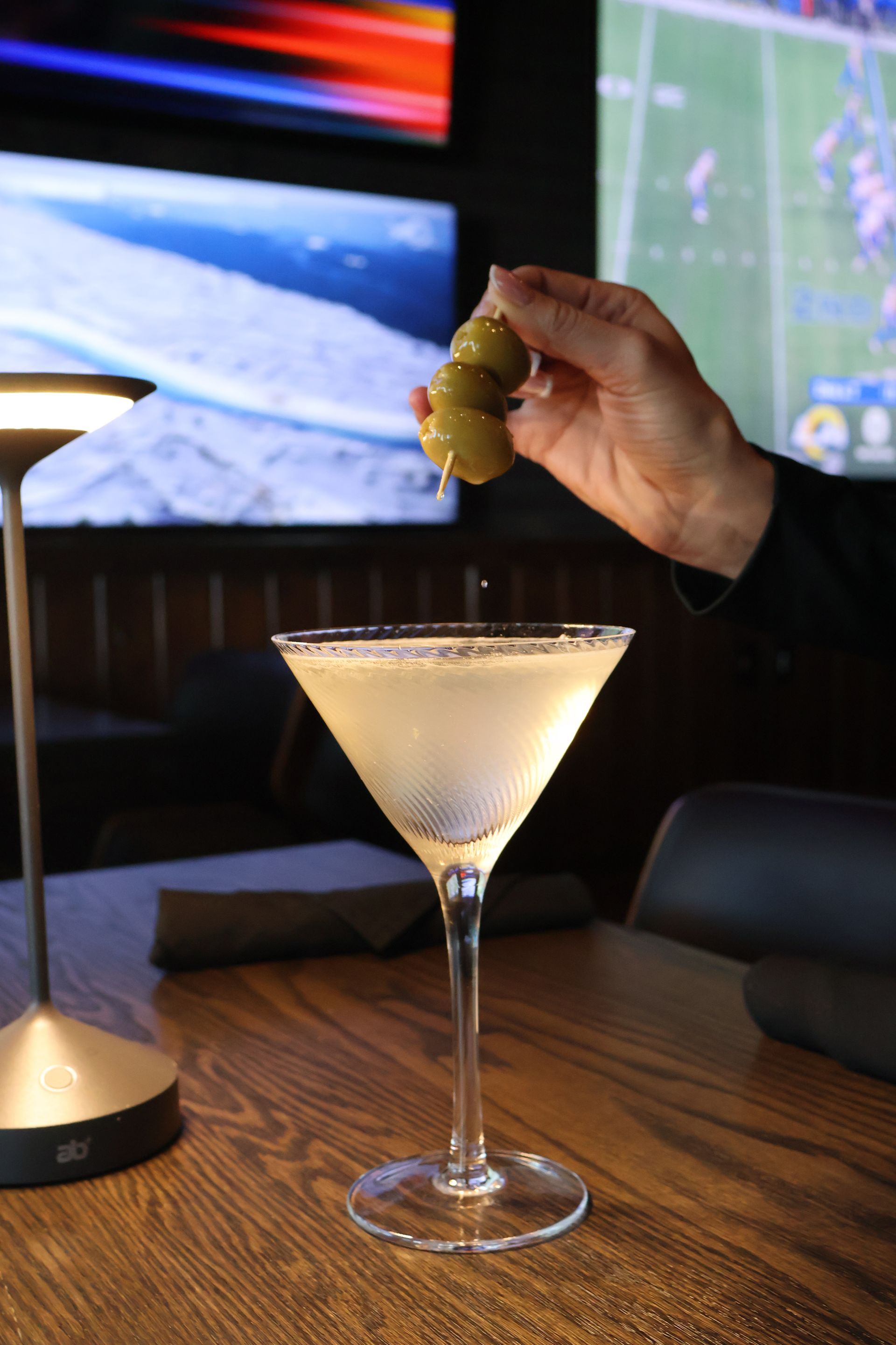 A hand drops a skewer of three olives into a martini glass on a wooden table in a dimly lit bar with television screens.