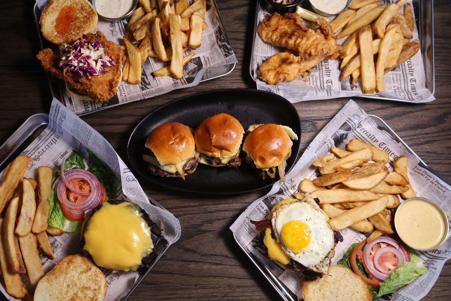 Four trays of pub-style food, including burgers, fish and chips, and sliders, arranged on a dark wooden table.